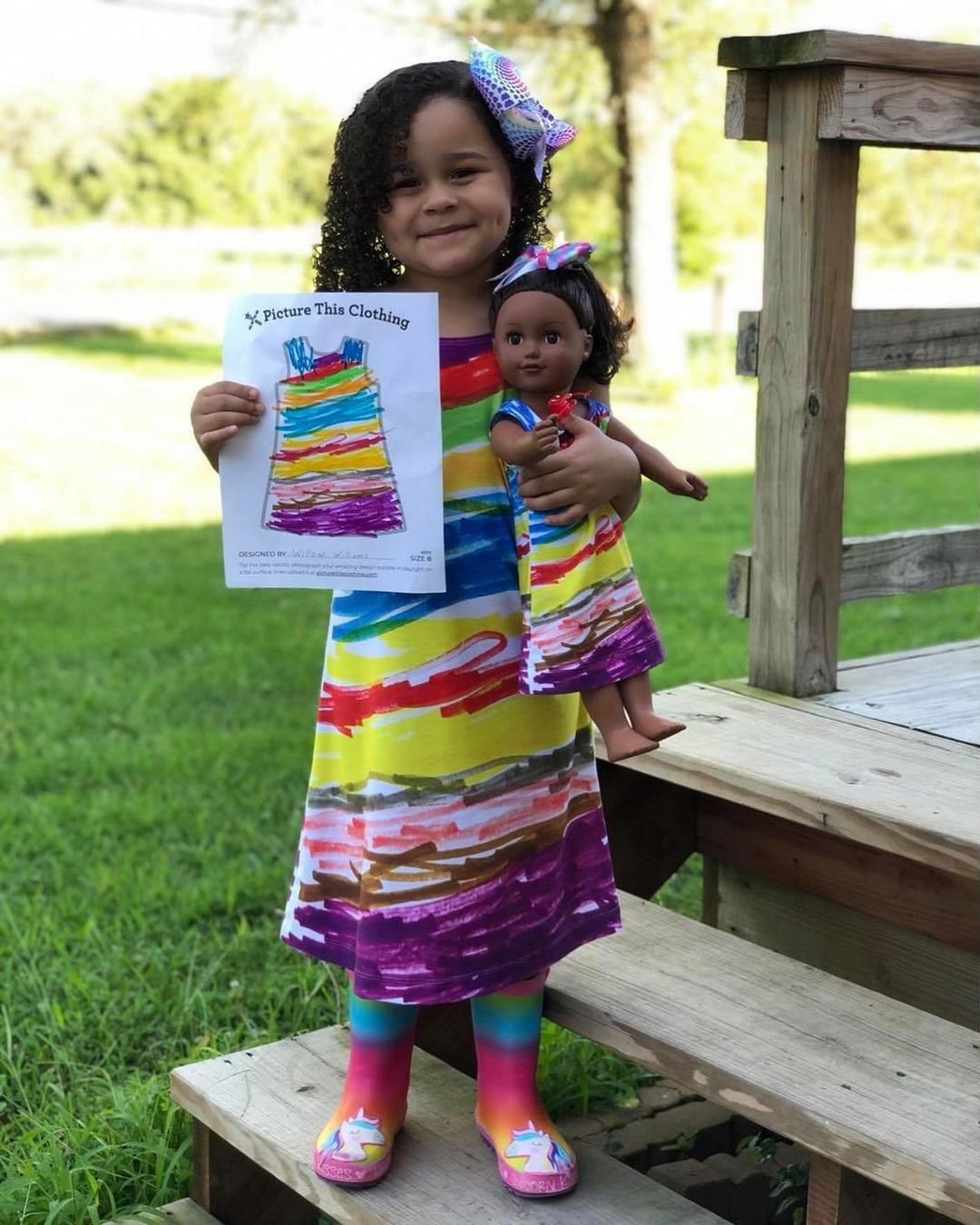Adorable little girl holding a paper with dress design, wearing the design she drew, and also holding a doll with a matching dress.