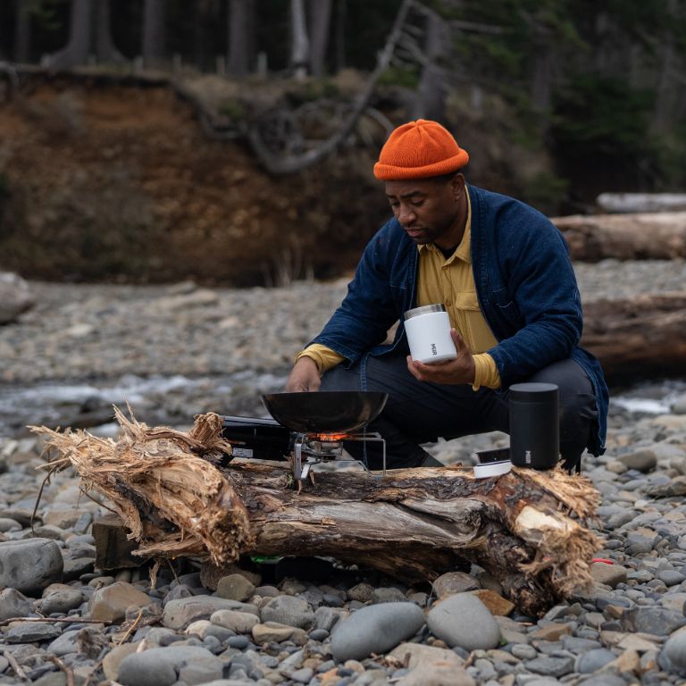 Man using the new white & black MiiR food containers to warm up food by the camp fire at Camp Yoshi