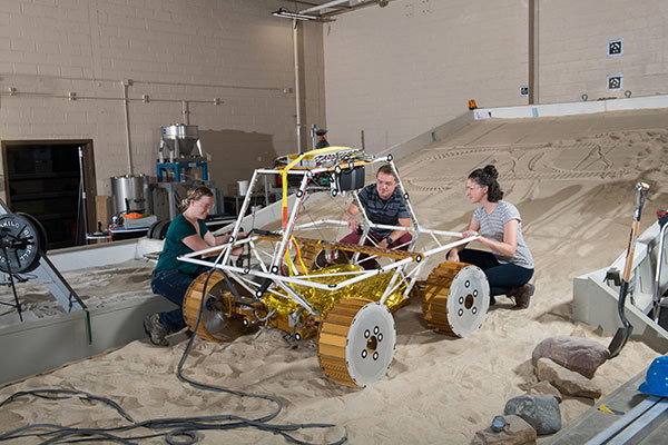 Three people crouch over a prototype rover that looks like a small dune buggy with golden wheels.
