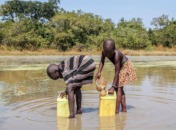Two children stand ankle-deep in a pond, filing large yellow vessels.