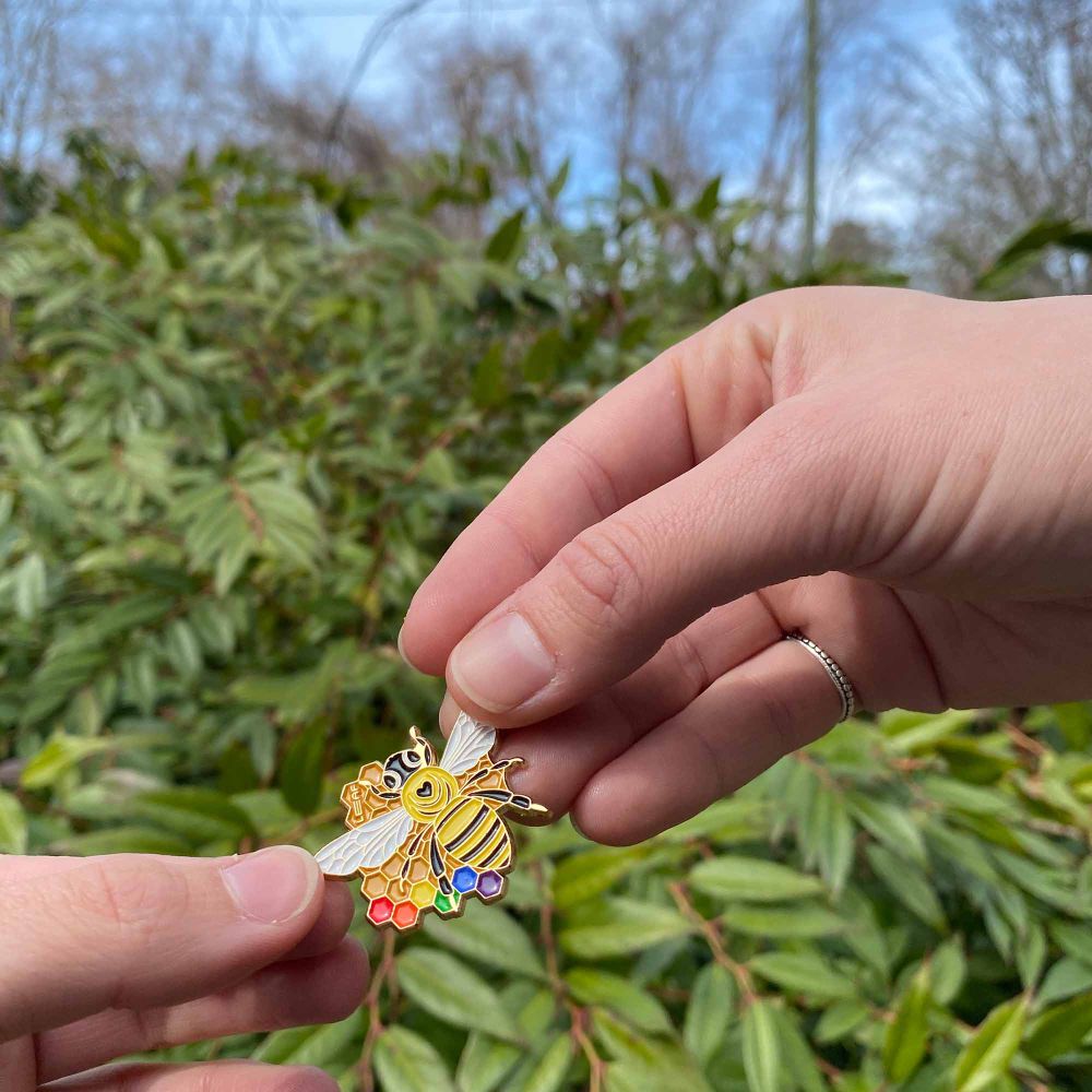 Enamel bee pin held in front of plants by two hands.
