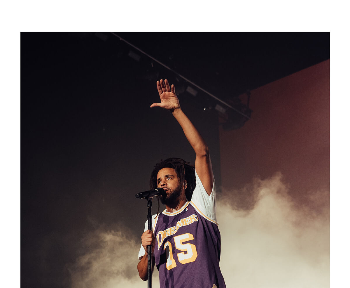 J.Cole holding a mic with his right hand and raising his left hand