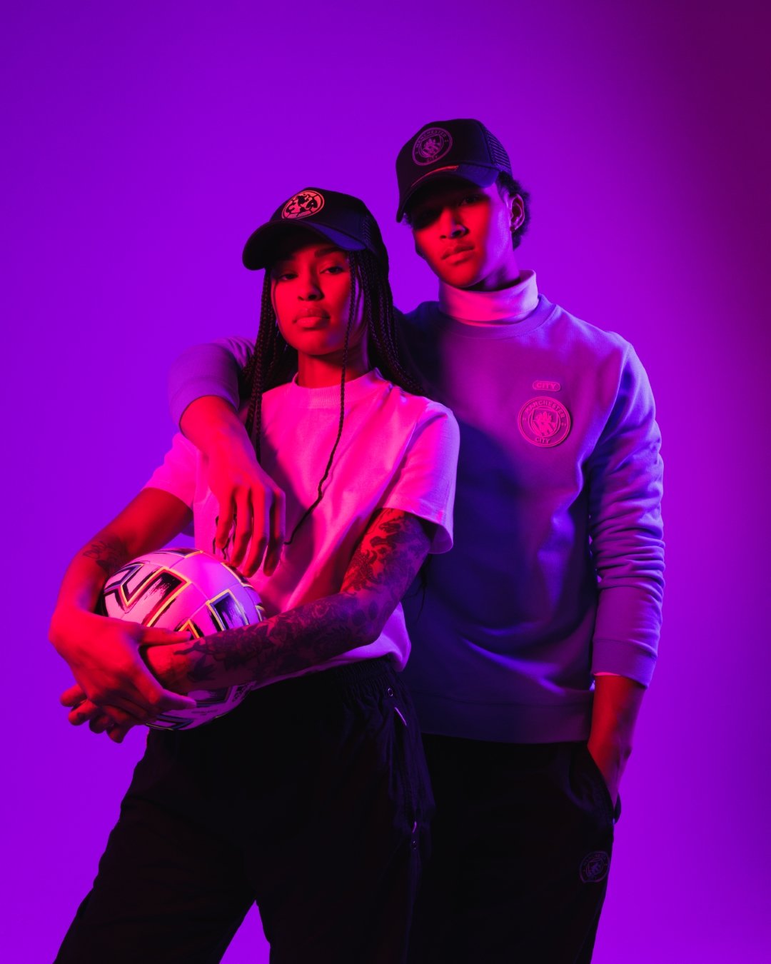 Woman and man pose in a purple-pink gradient environment. Women on left holds a football and wears the Club America Atmosphere Trucker. Man on right has his arm over the woman's shoulder, and wears the Manchester City Atmosphere Trucker.