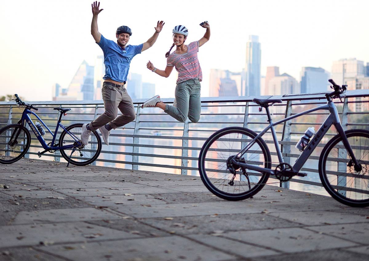Cyclists wearing helmets while jumping up in air with Trek-branded bikes, body of water, and cityscape in background