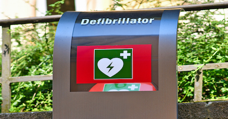 Defibrillators in einem Outdoorkasten, der mit der Aufschrift "Defibrillator" gekennzeichnet istVerschiedene Menschen bei einer Brandschutzhelfer- Ausbildung