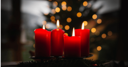 Adventskranz mit roten, brennenden Kerzen im Vordergrund, im Hintergrund ist ein  beleuchteter Weihnachtsbaum