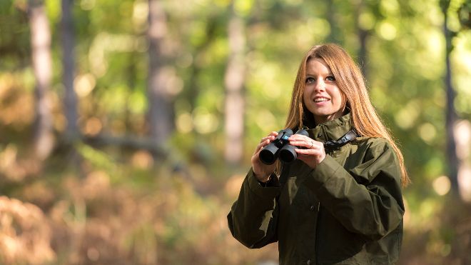 A young woman outside on a bright day, she has binoculars poised and eyes raised upwards ready to watch birds