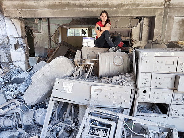 A wide angle photo of a destroyed physics laboratory and the scientist it once belonged to. The woman with long dark hair, wearing black jeans and a red t-shirt is the only spec of colour in the image as she sits on top of a large pile of grey rubble that contains stacks of now broken laboratory instruments. She looks exhausted, crossing her arms in a motion that looks almost like she’s hugging herself.