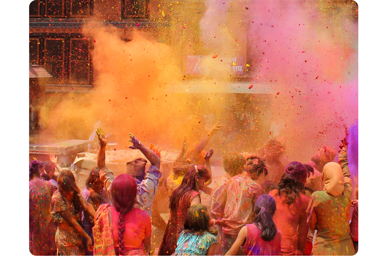 India’s Holi festival. A large group of people, mid-celebration, are seen from behind in the middle of a colorful cloud of pinks, oranges, and purples.