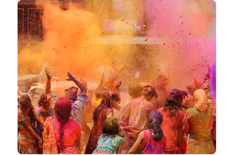 India’s Holi festival. A large group of people, mid-celebration, are seen from behind in the middle of a colorful cloud of pinks, oranges, and purples.