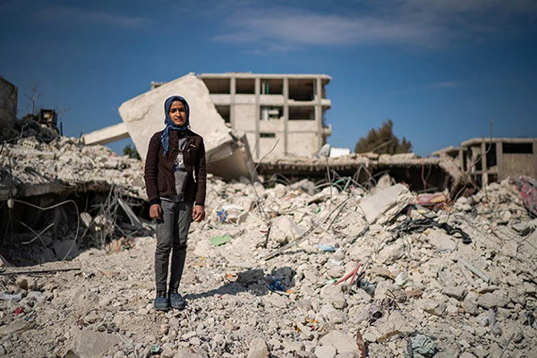 A photo of a girl in dark trousers, a grey T-shirt, a brown cardigan and a blue headscarf stands in front of a pile of concrete rubble. Her arms are hanging by her sides as she looks at the camera with a serious expression. Behind her, under a bright blue sky, are the remnants of a destroyed multi-storey building.