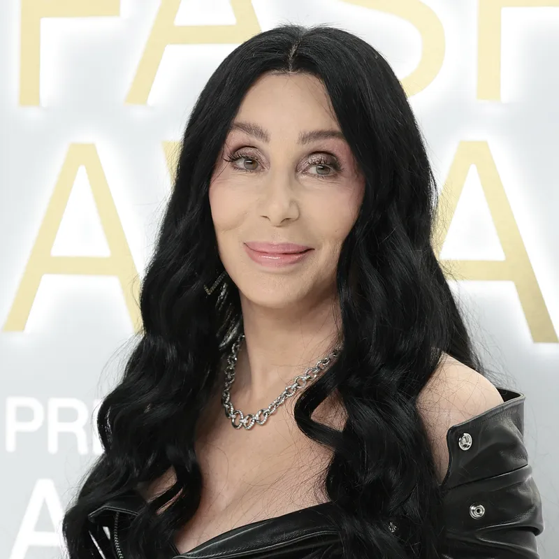 Cher appears at an event in a black off-the-shoulder motorcycle jacket with long black curls.