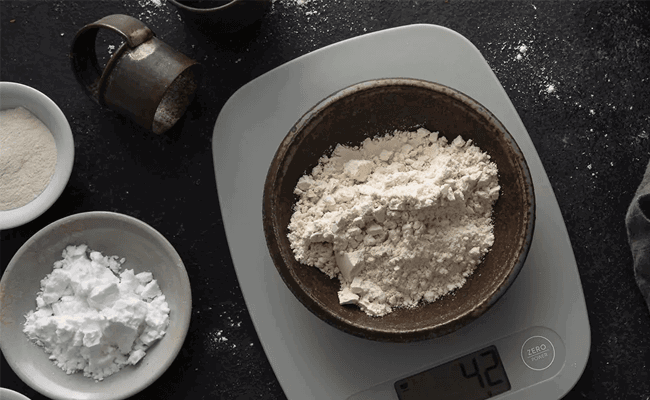 Oat flour in a bowl on a kitchen scale, beside a smaller bowl of potato starch, with a larger bowl of whole grain gluten-free flour off to one side