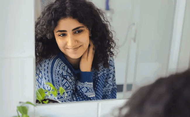 A young woman with curly hair looks into the bathroom mirror and smiles