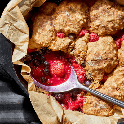 Bursting with Berries Cobbler in a cast iron skillet lined with parchment paper with a big metal spoon