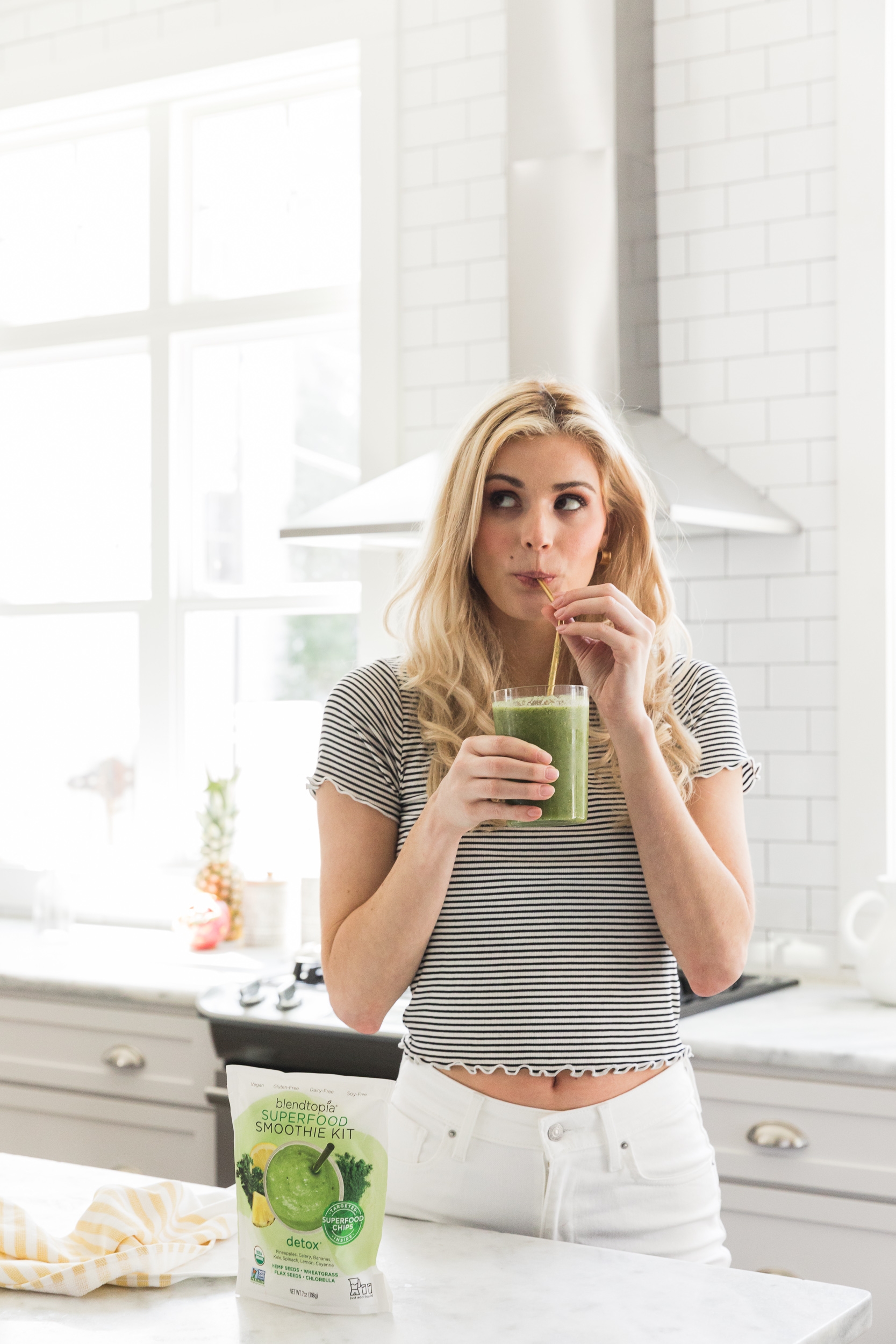 A blonde female in white jeans and a black & white striped short sleeve shirt holding and sipping a green smoothie.