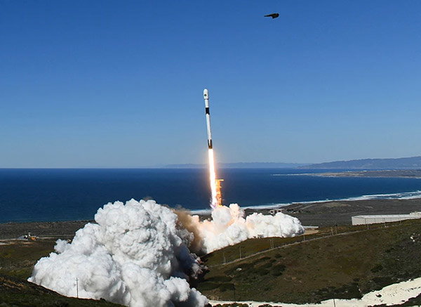 A slim rocket takes off from a spaceport next to the ocean, set amid green hills. Large white clouds of smoke billow out from the launch pad. It’s a beautiful sunny day, with blue skies.