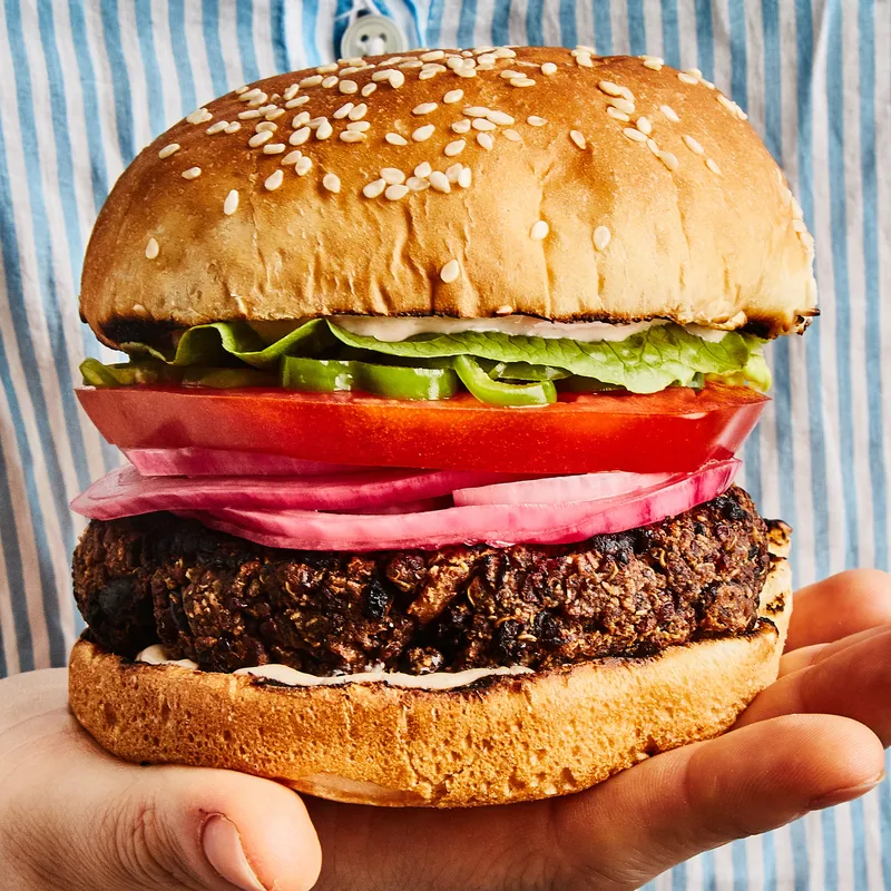 A hand holding a black bean veggie burger on a bun, topped with onions, tomato, lettuce, and pickles.
