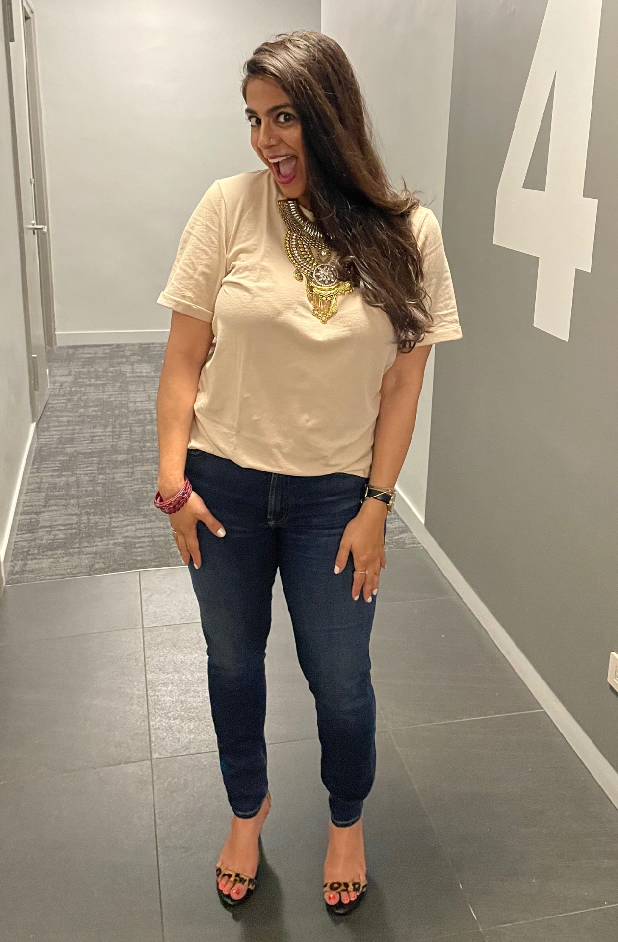 Woman wearing a beige tshirt. statement necklace and blue jeans making an excited face