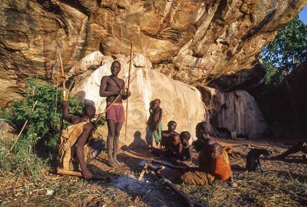 A group of people, including several children, sits on the dry ground around a campfire under a huge overhanging rockwall. Soft evening light casts shadows onto the rock. A small dog sits in the background. One man is standing, holding a spear, apparently conversing with another man who is crouching on the ground while holding a large bow.