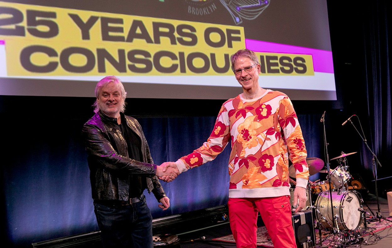 David Chalmers, dressed in a black leather jacket, shakes hands with Christof Koch, in a vibrantly coloured pullover, both smiling widely in front of a screen that reads “25 years of consciousness