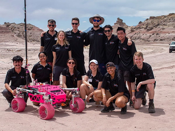 A group of young adults stand in the desert with their bright-pink prototype rover. 