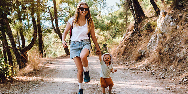 Photo of a woman smiling and holding her toddler's hand as they are walking an outdoor trail
