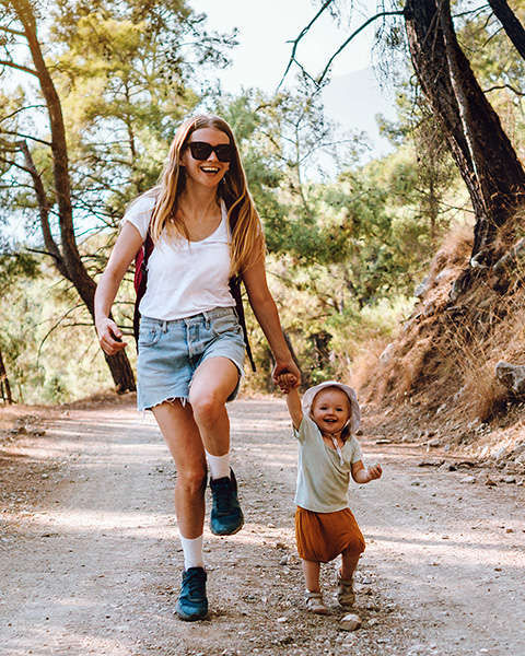 Photo of a woman smiling and holding her toddler's hand as they are walking an outdoor trail