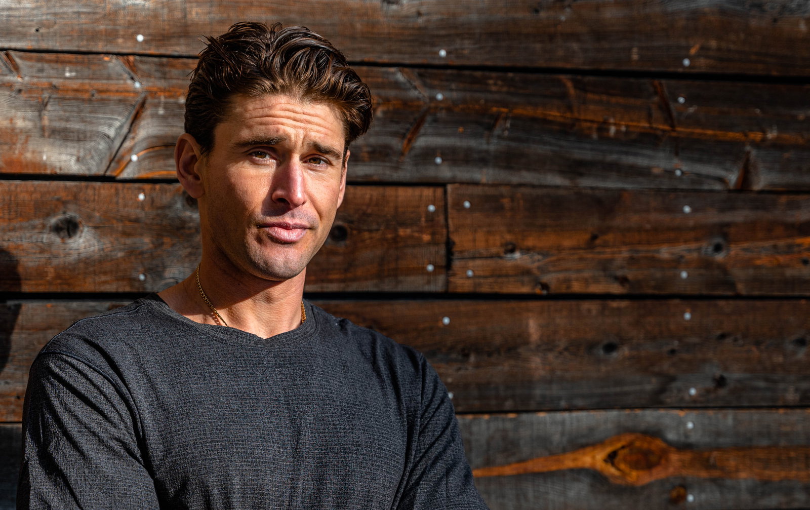 alt="Ben Greenfield stands in front of an old barn wall with the sun shining brightly on him"