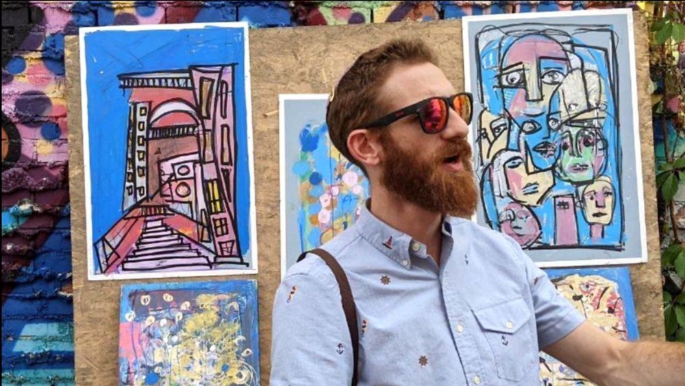 Benny Blindspots standing in front of a display of his paintings.