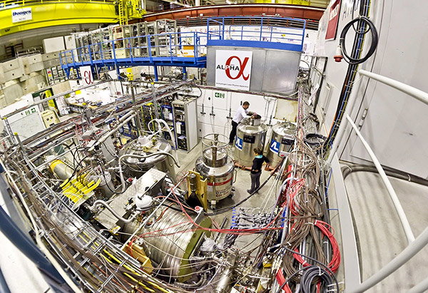 A wide-angle view of a room full of shiny metal machinery and control panels, connected by densely packed bunches of cables. A sign in the background identifies this as the ALPHA experiment. Two staff members are inspecting one of the cryotanks.