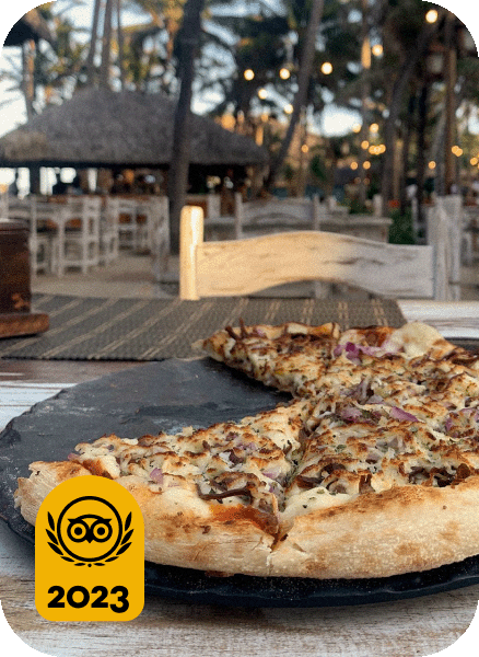 Image description: Whole pizza with a couple slices removed with palm trees in the background. Travelers’ Choice Best of the Best 2023 yellow logo in the bottom left corner.