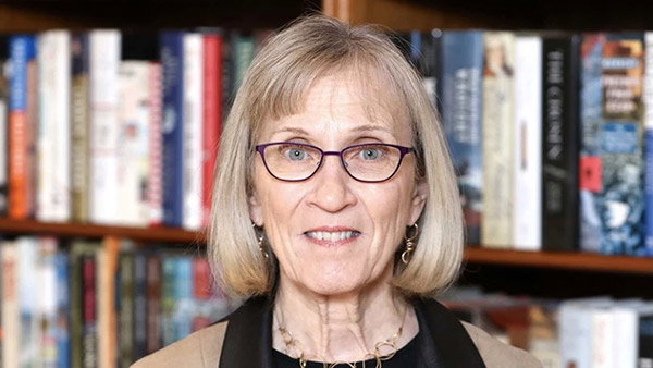 A portrait photo of Claudia Goldin, an older woman with chin-length light blonde hair, blue eyes and glasses. She’s smiling at the camera while standing in front of a bookcase.