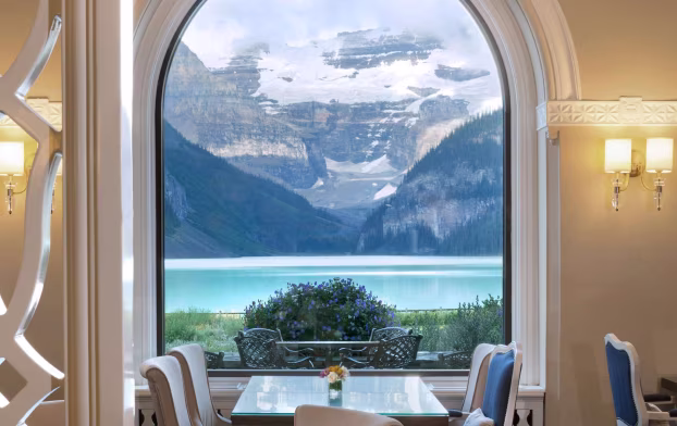 An empty dining room table with four chairs sits near a large open window revealing snow-capped mountain views and a glacier-fed lake.