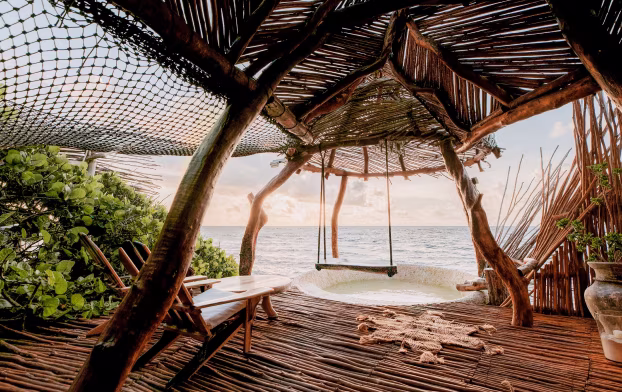 A hotel balcony constructed of wooden slats, tree trunks and fish netting is adorned with chairs, a swing and a hot tub, and overlooks the ocean.