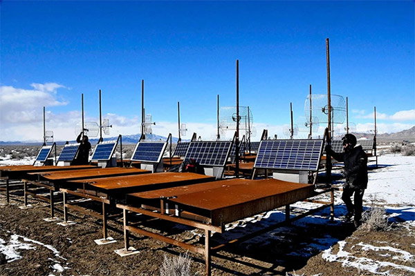 Under a bright blue sky, two people in dark winter clothing inspect rows of surface detectors that look like large rusty metallic tables powered by solar panels. A dusting of snow covers the ground.