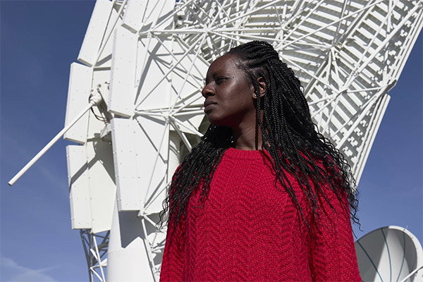 A person with long black black microbraided hair stands in front of a large white telescope dish. They look off into the distance as their knitted, bright-red jumper stands out sharply against the cloudless blue sky.