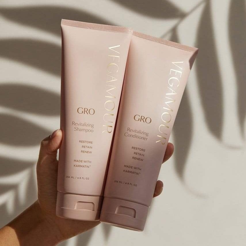 A hand holding the GRO Revitalizing Shampoo and Conditioner Kit from Vegamour with palm leaf shadows in the background.