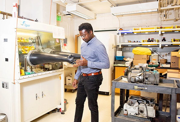 A man with short black hear and glasses, wearing black jeans and a blue shirt, stands inside a chemistry lab. He seems to shake hands with the black gloves sticking out of a glovebox, a type of sealed fumehood filled with inert gas. Next to him, two large vacuum pumps sit on a plastic trolley and crammed workbenches are visible in the background.