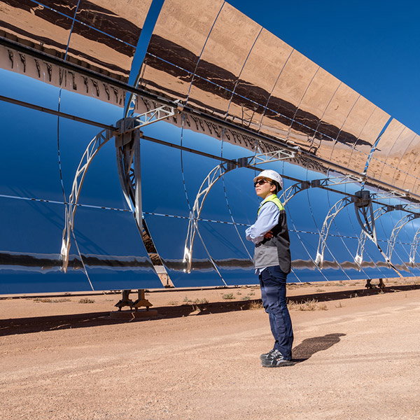 A woman wearing a white hard hat, sunglasses, and hi-vis jacket stands in front of a long array of parabolic mirrors, against a backdrop of blue sky