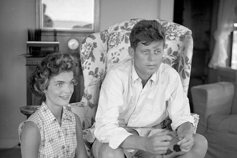 black and white photo of John F. Kennedy sitting on chair, Jackie Bouvier to left sitting on floor, at Kennedy compound 