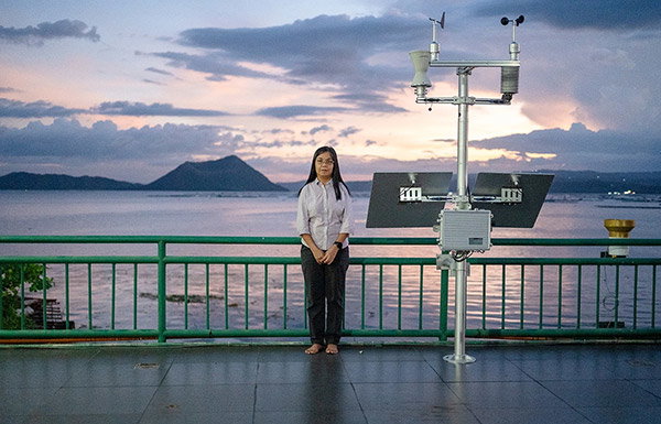 A barefoot woman with black shoulder-length hair, wearing a white shirt and dark trousers, stands next to a monitoring station, overlooking Taal volcano in the far distance. She is standing in front of a green railing, with a calm sea and darkening sky behind her. 