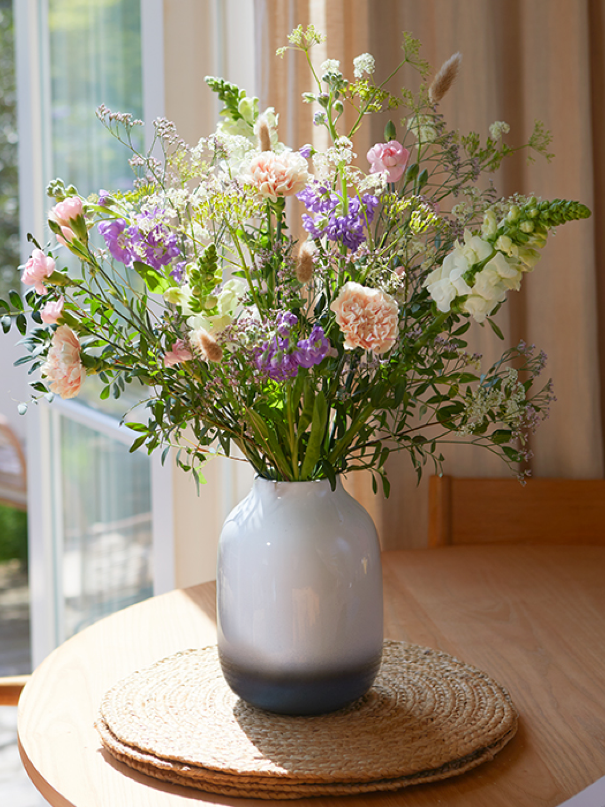 Ein heiterer Blumenstrauß mit filigranen Blüten in zarten Pastelltönen steht in einer Keramikvase auf dem Esstisch.