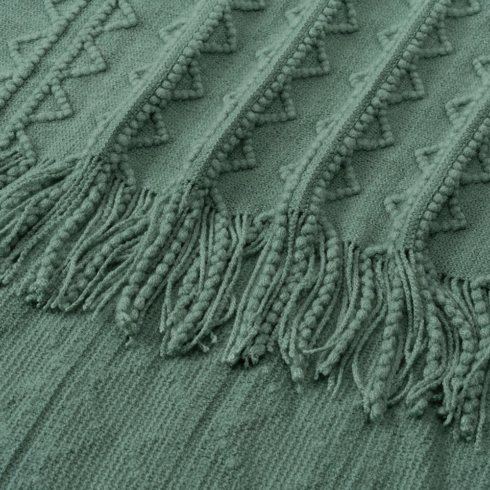 A close-up image of a green blanket with a knitted pattern and tassels on the edge.
