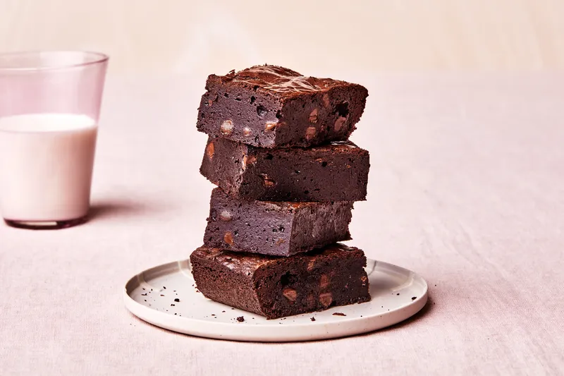 Triple Chocolate Brownies stacked on a white plate with a glass of milk behind it. 