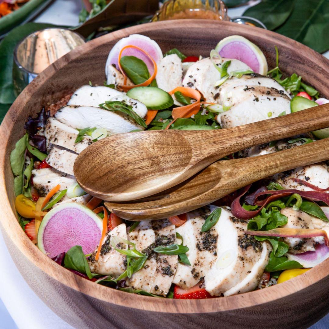 a large wooden salad bowl with chicken and a variety of vegetables, plus two wooden serving spoons
