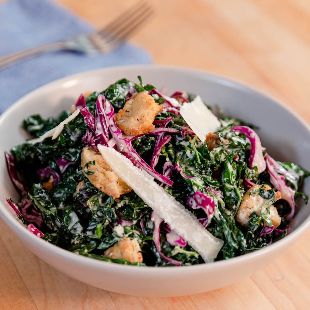 a white ceramic bowl containing a kale salad with cabbage, croutons, and parmesan