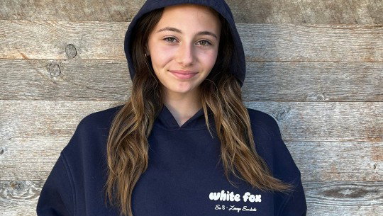 A teen girl smiles in a blue hooded sweatshirt with the White Fox logo