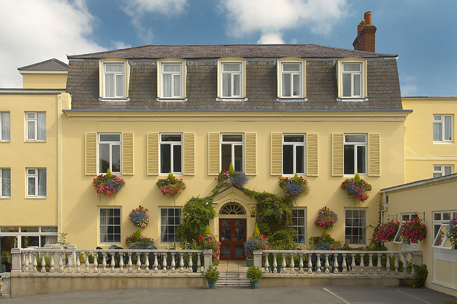 The front entrance of Les Rocquettes Hotelon a summers day, the building a light creamy colour with blooming flowers outside.