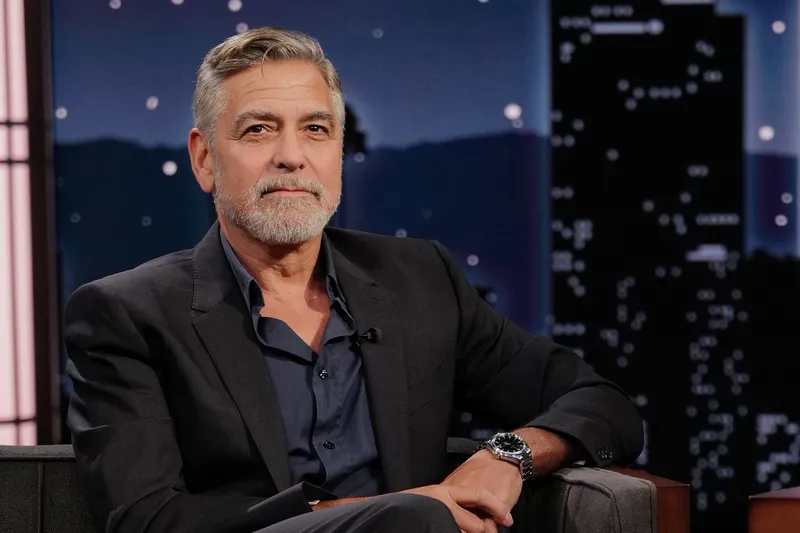 George Clooney sitting on grey cushioned armchair with legs crossed during late night TV talk show interview; cityscape backdrop